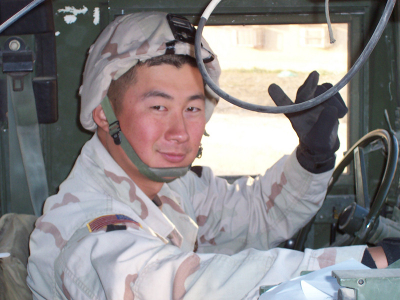 Staff Sergeant Kyu Hyuk Chay in the driver's seat of a Humvee in Afghanistan, wearing desert camouflage and a helmet, looking toward the camera.